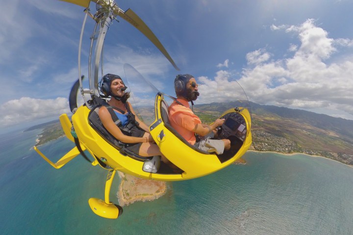 Two people flying a yellow gyrocopter over a coastal landscape with blue sky.