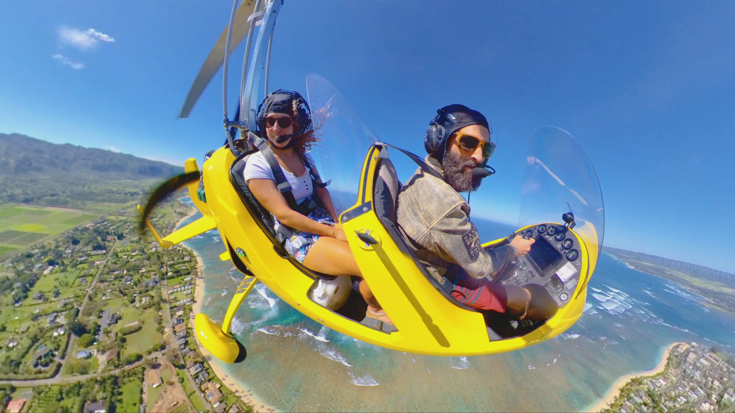 Two people flying in a yellow gyrocopter over a coastal landscape on a clear day.