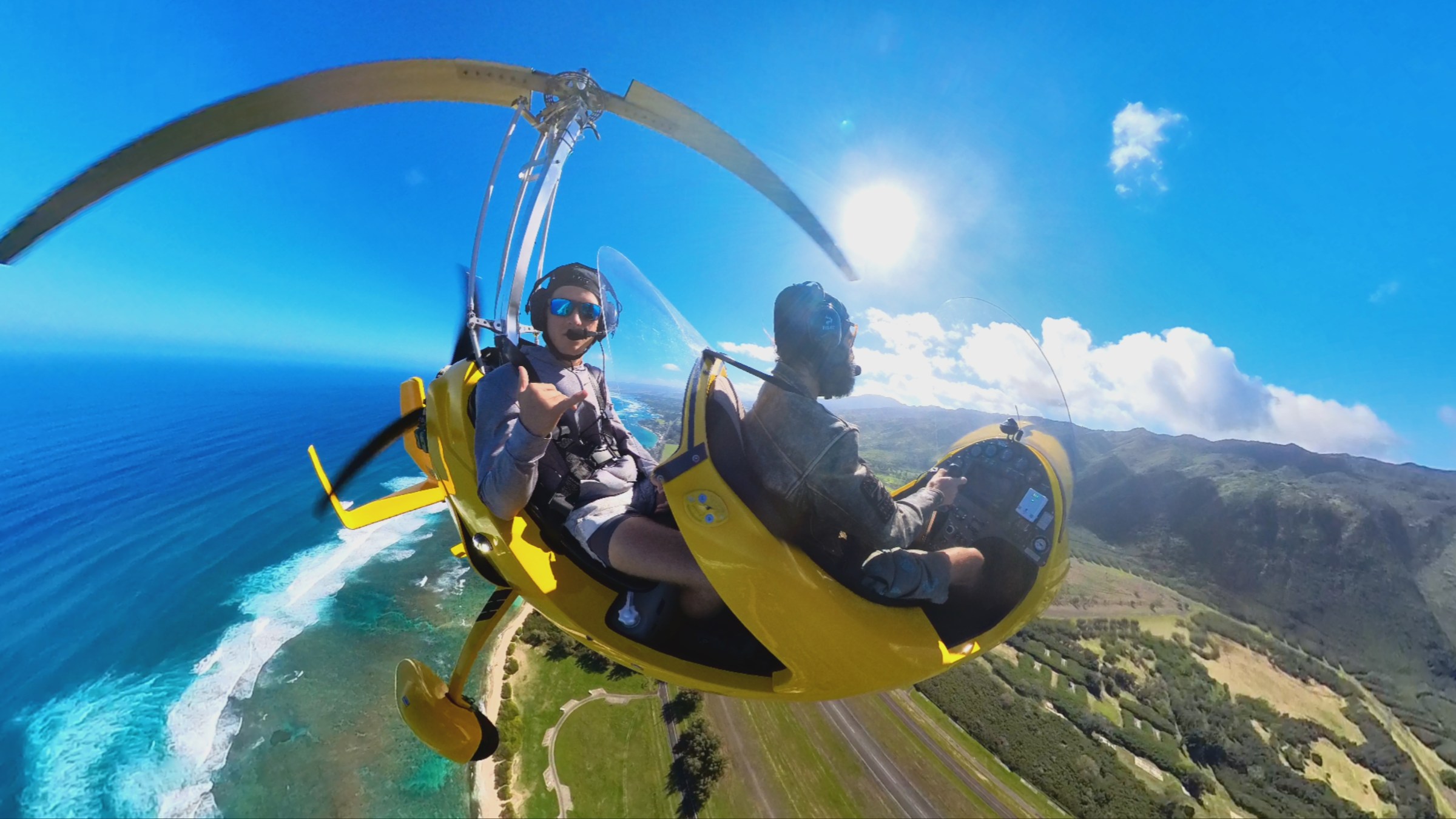 Two people flying a yellow gyrocopter over a coastal landscape with clear skies.