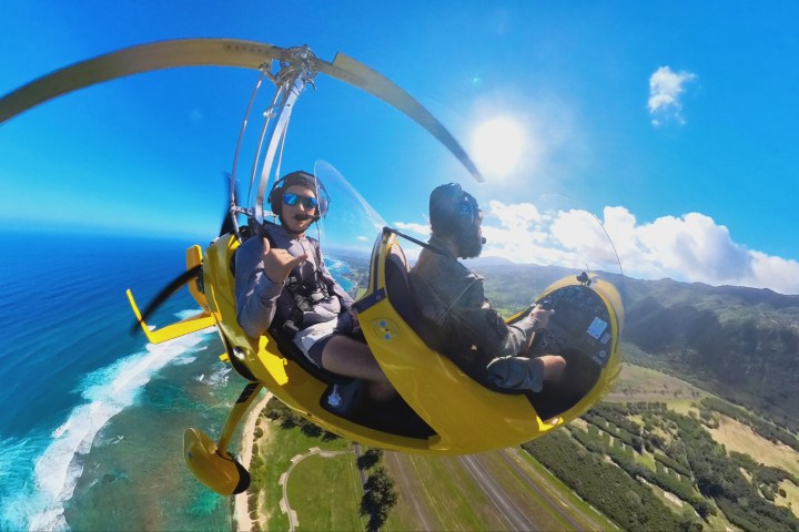 Two people flying a yellow gyrocopter over a coastal landscape with clear skies.