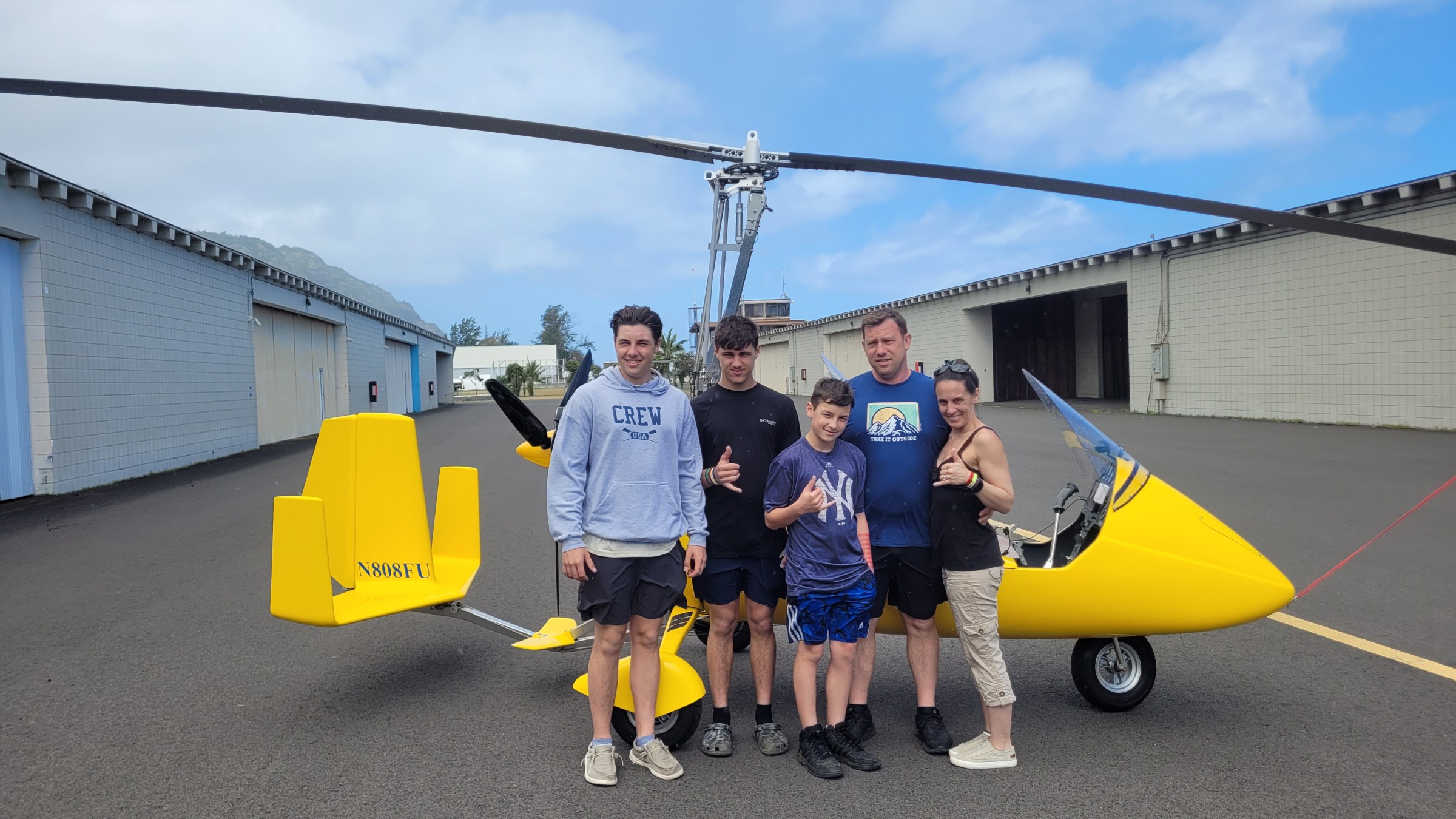 Group of people standing in front of a yellow gyroplane on a runway with hangars in the background.