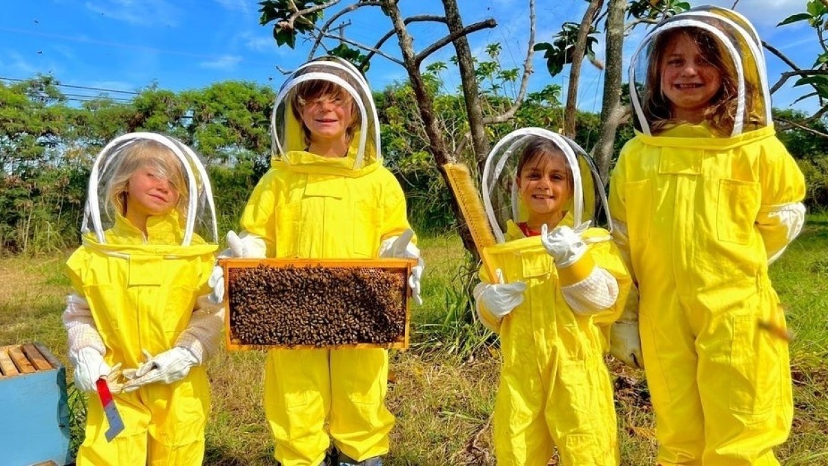 Four children in yellow beekeeping suits holding honeycomb frames in a sunny garden.