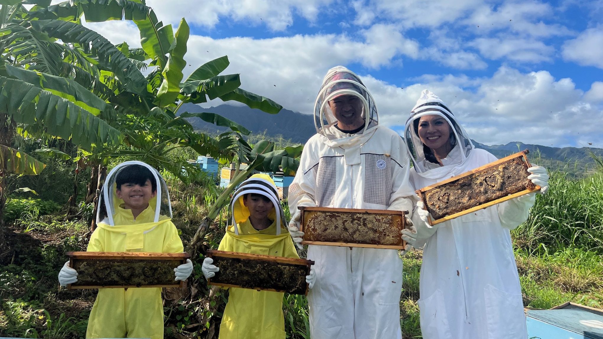Group of people in beekeeper suits holding honeycombs in a farm setting with banana trees.