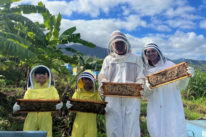 Group of people in beekeeper suits holding honeycombs in a farm setting with banana trees.
