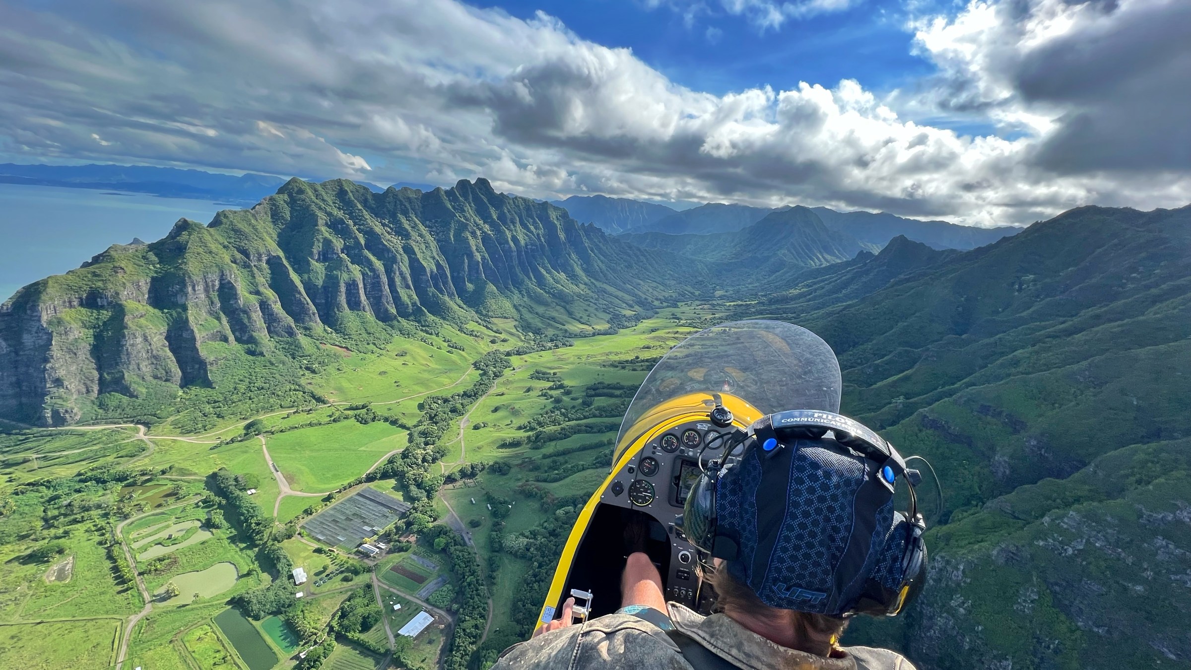 Person flying in open cockpit plane over lush, mountainous landscape with ocean view.