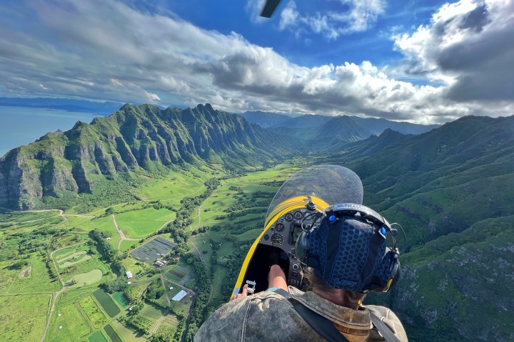 Person flying in open cockpit plane over lush, mountainous landscape with ocean view.