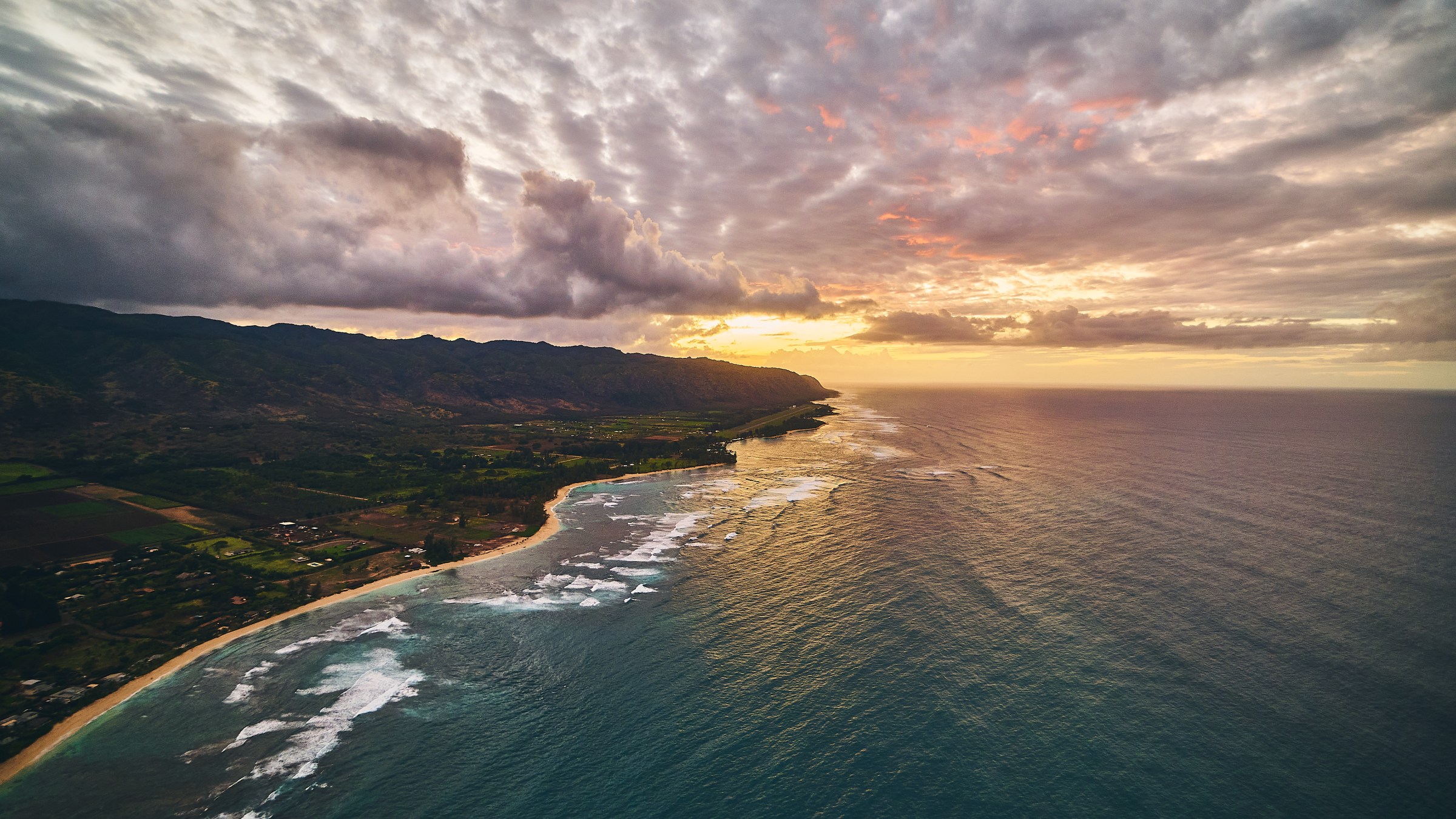 Sunset over a coastline with clouds, ocean, and distant mountains.