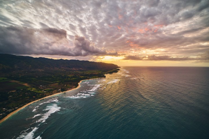 Sunset over a coastline with clouds, ocean, and distant mountains.