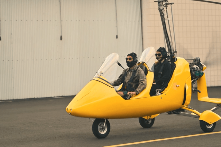 Two people in a yellow autogyro parked on an airport tarmac.