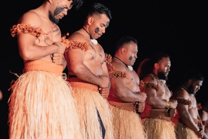 Men in traditional attire with face paint and grass skirts, standing on stage at night, arms crossed.