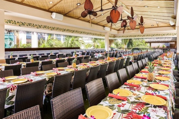 Long tables with floral cloths and yellow plates set for dining under a tropical pavilion.