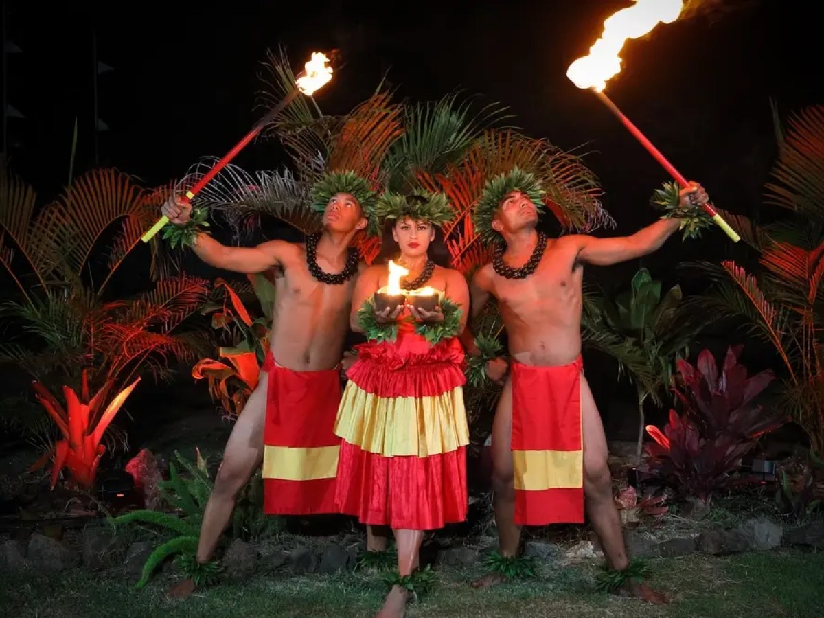 Three dancers in traditional attire holding torches, surrounded by tropical plants at night.