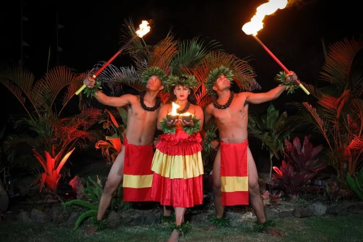 Three dancers in traditional attire holding torches, surrounded by tropical plants at night.