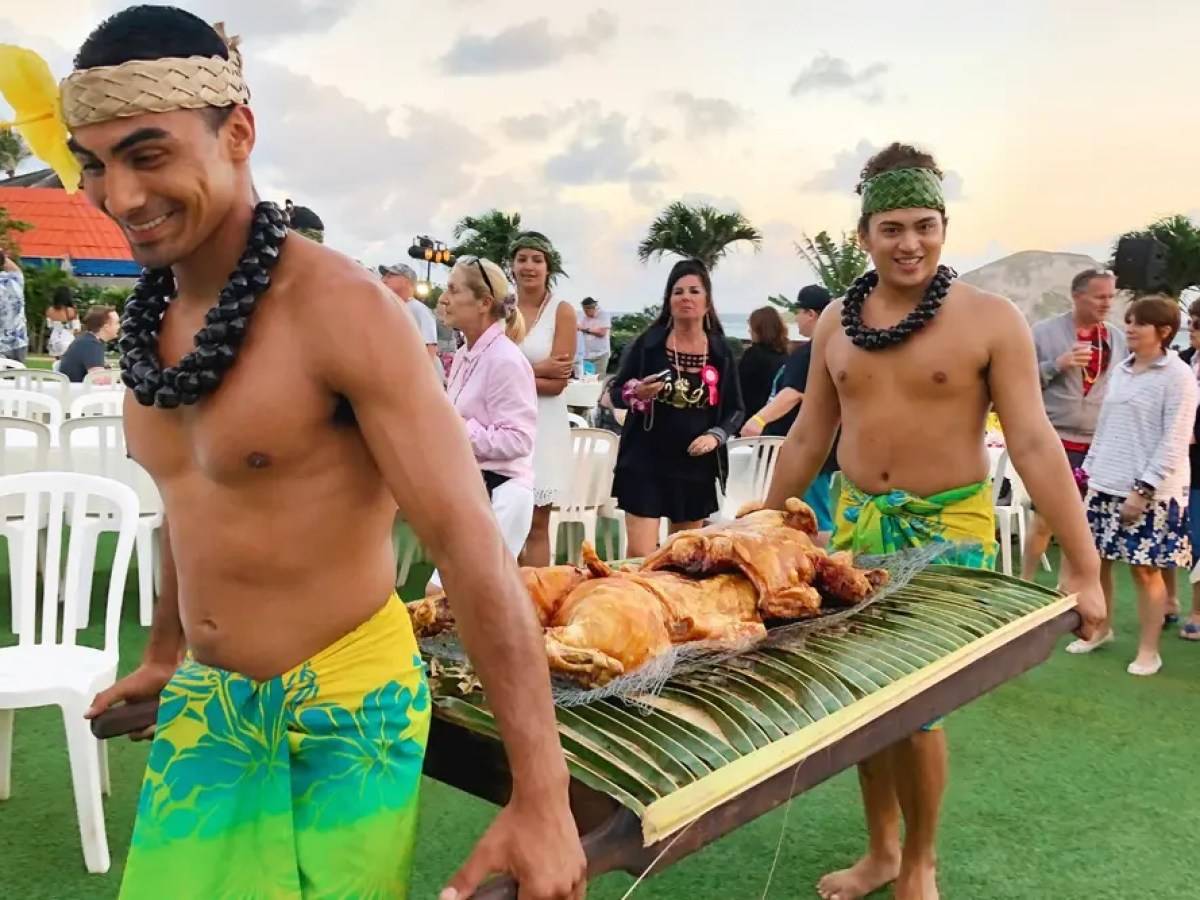 Two men in traditional attire carry a tray with roasted meat at an outdoor event.