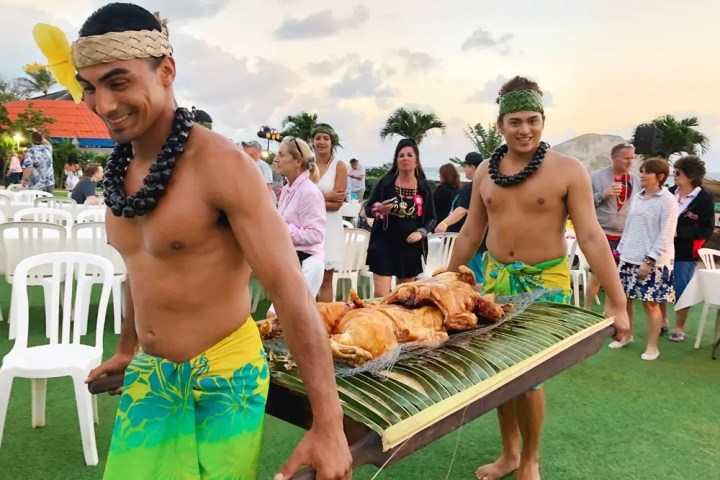 Two men in traditional attire carry a tray with roasted meat at an outdoor event.