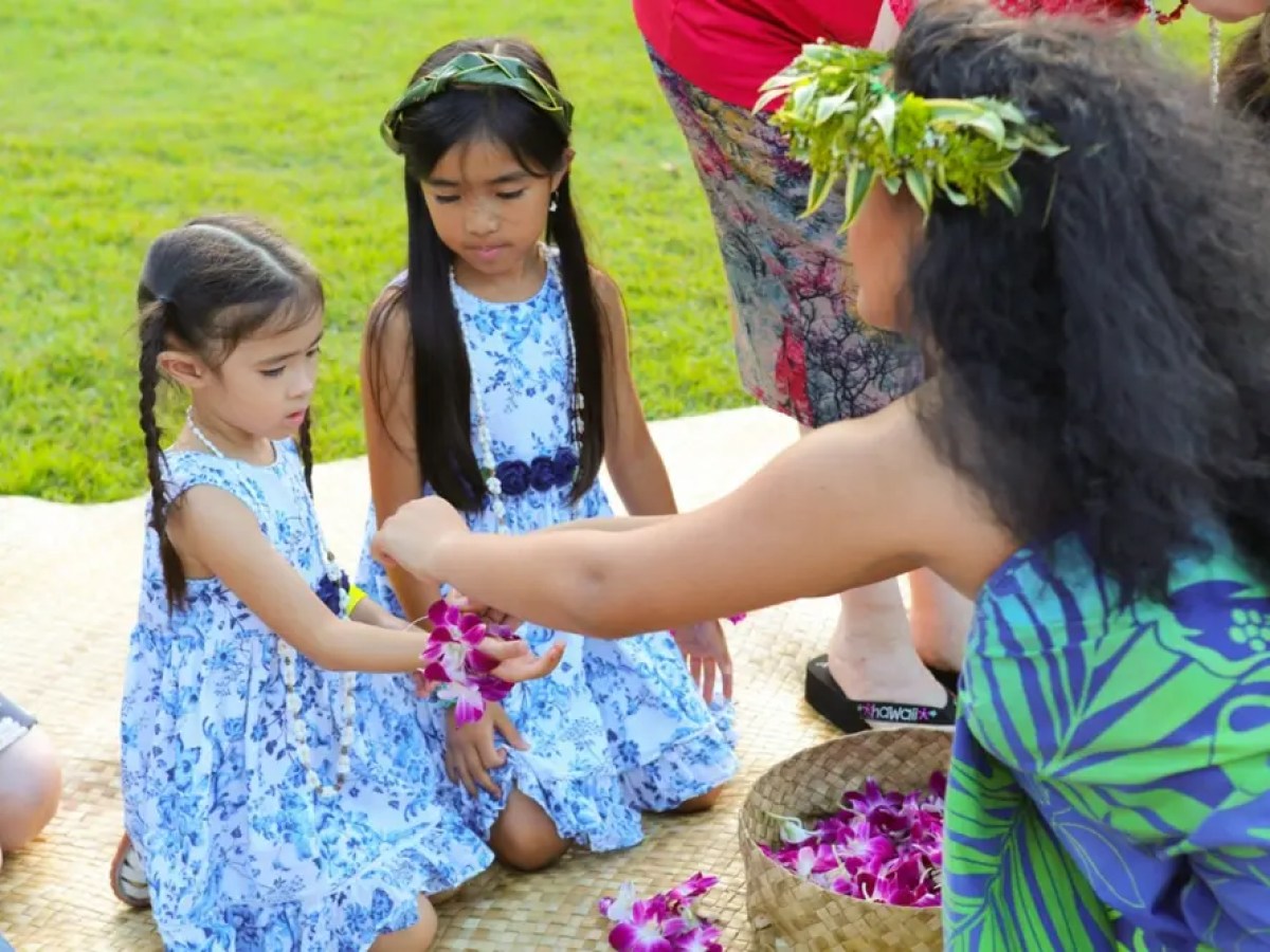 Woman in floral dress and crown gives leis to two girls in blue dresses sitting on grass mat.