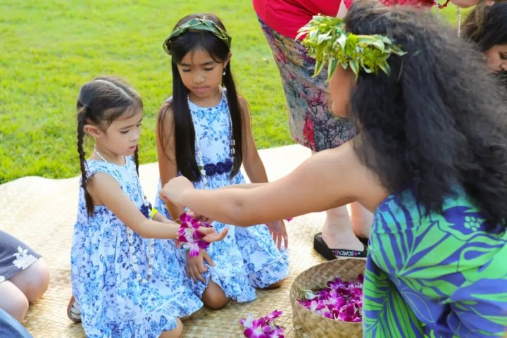 Woman in floral dress and crown gives leis to two girls in blue dresses sitting on grass mat.