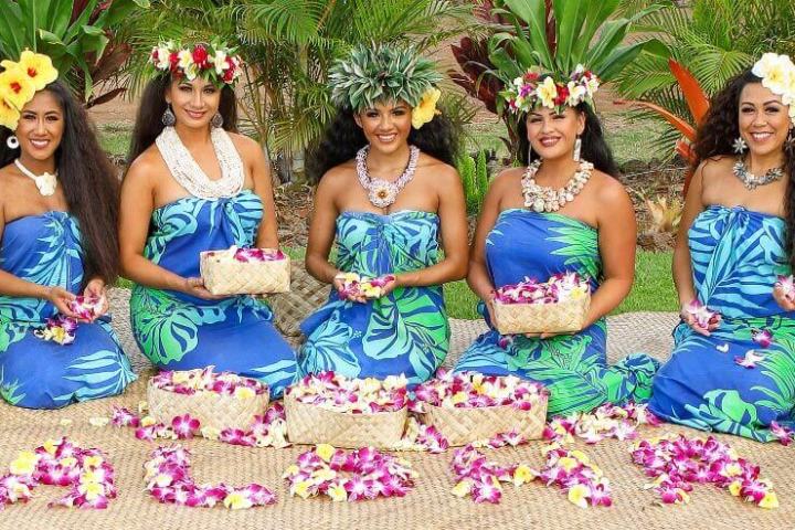 Seven women in blue dresses with flower crowns holding baskets of petals, spelling 'Aloha' on ground.