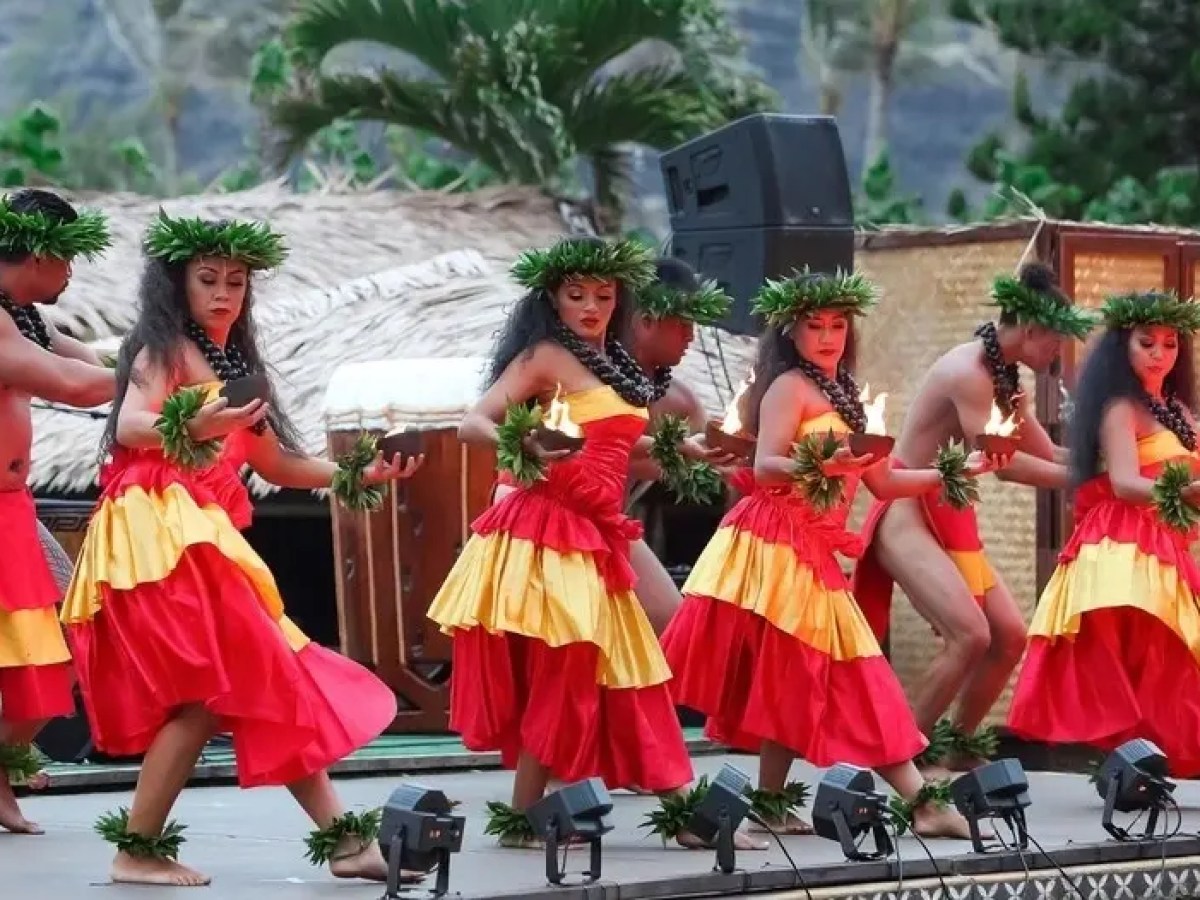 Six dancers in red and yellow costumes performing a traditional hula dance with torches on a stage.