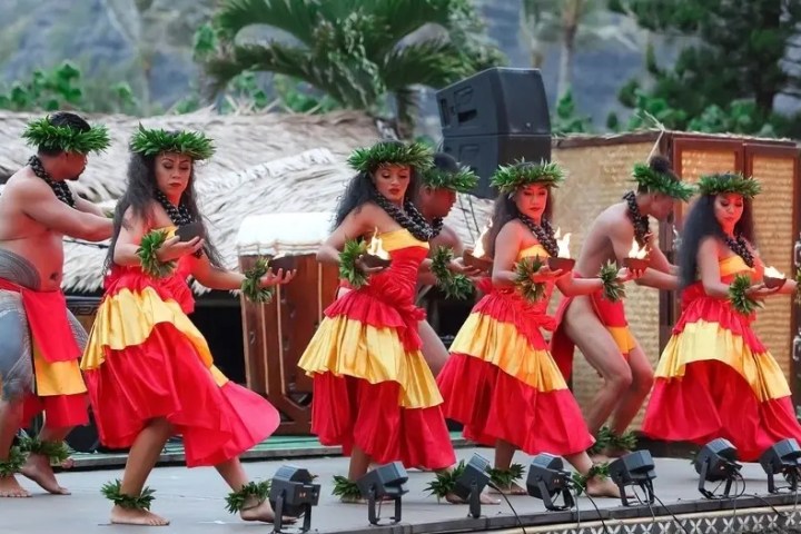 Six dancers in red and yellow costumes performing a traditional hula dance with torches on a stage.