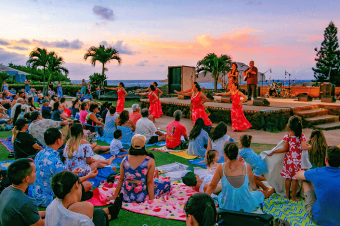 Crowd watching outdoor dancers on stage at sunset, surrounded by palm trees.