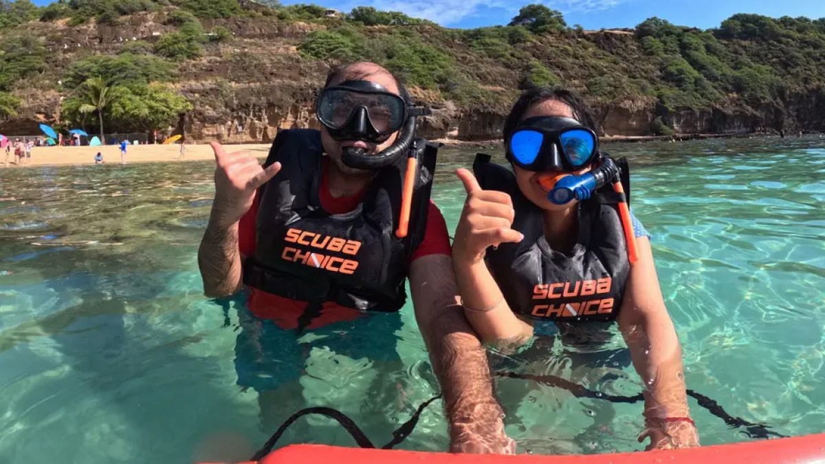 Two people in scuba gear, giving thumbs up in clear water near a beach.