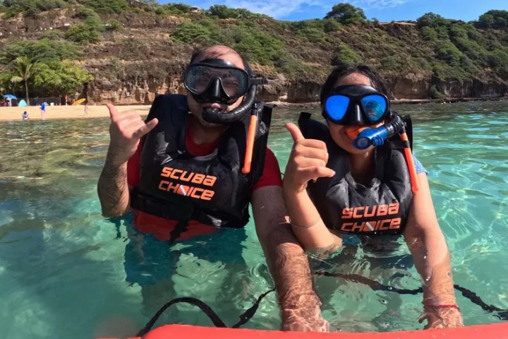 Two people in scuba gear, giving thumbs up in clear water near a beach.